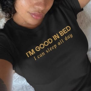 Puede incluir: Camiseta negra con el texto dorado "I'M GOOD IN BED I can sleep all day". La persona que lleva la camiseta tiene el pelo rizado oscuro y pecas.