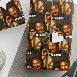 May include: Gift wrapping paper featuring a repeating pattern of a man's face with the text "IT IS A GIFT!". The wrapped gifts are tied with twine and adorned with white tags. A ball of twine and scissors are also visible.