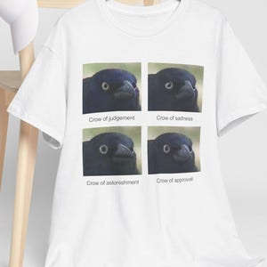 Puede incluir: Camiseta blanca con una imagen de cuatro paneles de un cuervo que muestra diferentes emociones. Los subtítulos dicen: "Crow of judgement", "Crow of sadness", "Crow of astonishment" y "Crow of approval". Una gorra de béisbol blanca está en una silla de madera.