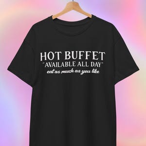 以下が含まれることがあります： 白文字で「HOT BUFFET」と書かれた黒いTシャツ。その下に「AVAILABLE ALL DAY」と「eat as much as you like」の文字。木製のハンガーにかかっています。