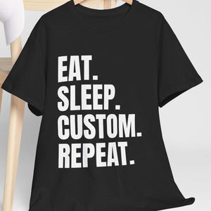 Op de afbeelding: Zwart T-shirt met de witte tekst "EAT. SLEEP. CUSTOM. REPEAT." aan een houten hanger. Een witte baseballpet rust op de rug van een lichte houten stoel.