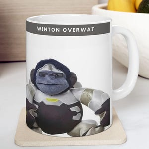 May include: White ceramic mug with a cartoon gorilla character in a futuristic outfit. The mug has the text "WINTON OVERWAT" printed on the top. The gorilla is primarily black, grey, and silver.