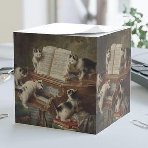 May include: A cube-shaped notepad with a vintage painting of kittens around a piano. The artwork shows black and white kittens studying sheet music on a dark wood piano. The scene has a classic, artistic feel.