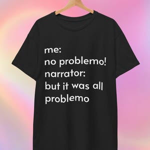 May include: Black t-shirt with the text "me: no problemo! narrator: but it was all problemo" in white. The shirt is hanging on a wooden hanger against a colourful, blurred background.