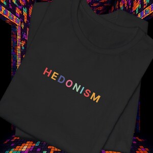 Puede incluir: Camiseta negra con la palabra "HEDONISM" bordada en colores arcoíris. La camiseta está doblada y se muestra sobre un fondo colorido y pixelado, creando una presentación vibrante y llamativa. Una prenda de vestir informal.