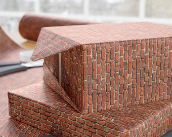 May include: Two rectangular boxes wrapped in paper featuring a realistic red brick pattern. The paper displays a repeating texture of rectangular bricks with dark mortar lines. A pair of black scissors and a black ribbon are placed nearby.