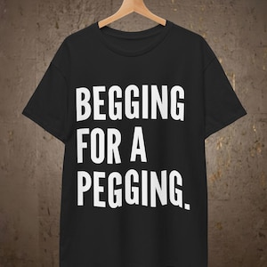 May include: A black short-sleeved t-shirt featuring the white text BEGGING FOR A PEGGING. printed in a bold, sans-serif font across the chest. The garment is displayed on a wooden hanger suspended from a metal rod against a textured wall.