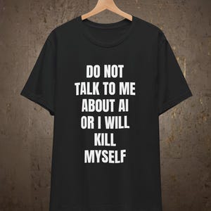 Może przedstawiać: Czarny t-shirt z krótkim rękawem z białym nadrukiem na przodzie o treści: DO NOT TALK TO ME ABOUT AI OR I WILL KILL MYSELF. Odzież jest zawieszona na drewnianym wieszaku na tle o fakturze w kolorze ciemnobrązowym.