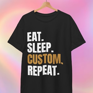 May include: Black t-shirt with the words "EAT. SLEEP. CUSTOM. REPEAT." in white and gold lettering. The shirt is hanging on a wooden hanger against a colourful, blurred background.