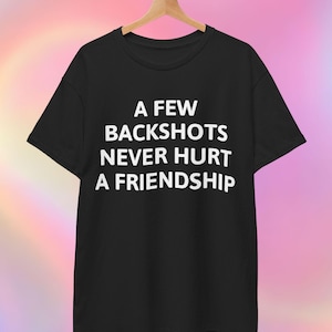 May include: Black t-shirt with the white text "A FEW BACKSHOTS NEVER HURT A FRIENDSHIP." The shirt is hanging on a wooden hanger against a colorful, blurred background. The t-shirt is a classic crew neck style.