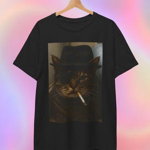 Può includere: T-shirt nero con l'immagine di un gatto con un fedora e una benda sull'occhio, con una sigaretta in bocca. Il gatto è al centro dell'immagine, su uno sfondo neutro.