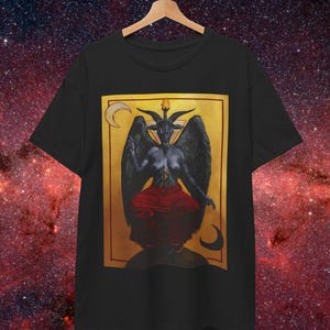 Pode incluir: Uma t-shirt preta de manga curta com uma impressão gráfica retangular de uma figura alada e com chifres de cabra, sentada numa posição de meditação. A figura veste uma peça vermelha sobre um fundo dourado com luas crescentes.