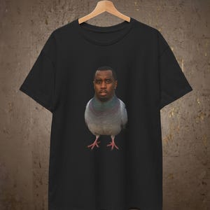 Op de afbeelding: Een zwart t-shirt met korte mouwen en een grafische print op de borst. Het ontwerp toont een duif met het hoofd van een man, met een realistisch vogel lichaam met grijze en iriserende nekveren en een menselijk gezicht met een serieuze blik.