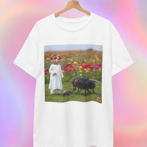 Könnte beinhalten: Weißes T-Shirt mit einem surrealen Bild eines Kindes in weißer Kleidung auf einem Blumenfeld, mit einer großen Fliege und einer kleineren. Das T-Shirt hängt an einem Holzbügel.