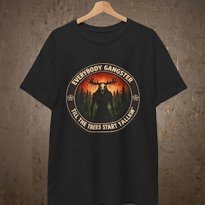 Op de afbeelding: Zwart t-shirt met een cirkelvormige afbeelding met de tekst "EVERYBODY GANGSTER TILL THE TREES START TALKIN'". De afbeelding toont een boslandschap met een wezen en geweien. Het t-shirt hangt aan een houten hanger.