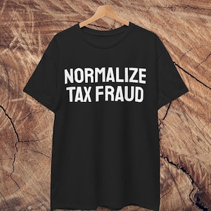 Könnte beinhalten: Schwarzes T-Shirt mit dem weißen Text "NORMALIZE TAX FRAUD". Das T-Shirt hängt an einem Holzbügel vor einem Holzmaserungshintergrund. Der Text ist in einer fetten, serifenlosen Schrift.