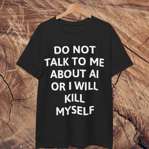 Puede incluir: Camiseta negra con el texto blanco "DO NOT TALK TO ME ABOUT AI OR I WILL KILL MYSELF". La camiseta está colgada en una percha de madera sobre un fondo de vetas de madera.