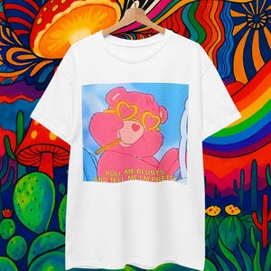 May include: White t-shirt featuring a pink cartoon bear wearing heart-shaped sunglasses and smoking a cigarette. The text "ROLL ME BLUNTS, AND TELL ME I'M PRETTY" is below the bear. The shirt is hanging on a wooden hanger against a colorful background.