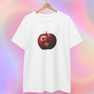 Könnte beinhalten: Weißes T-Shirt mit einer skurrilen Grafik eines roten Apfels mit Gesicht, der die Zunge herausstreckt. Das Shirt hängt an einem Holzkleiderbügel vor einem bunten, verschwommenen Hintergrund. Das Design ist verspielt und auffällig.