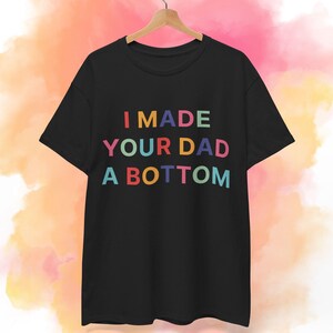 Könnte beinhalten: Schwarzes T-Shirt mit dem Text "I MADE YOUR DAD A BOTTOM" in bunten Buchstaben. Das Shirt hängt an einem Holzbügel vor einem farbenfrohen, abstrakten Hintergrund.