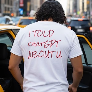 May include: A white t-shirt with the text "I told ChatGPT about u." in red lettering. The person is standing next to a yellow taxi cab on a city street with tall buildings in the background.