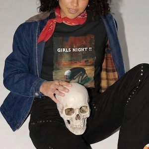 May include: Black t-shirt with the text "GIRLS NIGHT !!" and a graphic of a person kneeling. The person is holding a white skull. The person is wearing a denim jacket and black pants.