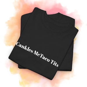 May include: Two black t-shirts are stacked on top of each other. The top shirt has the words "Cankles McTaco Tits" printed in white, diagonally across the front. The background has a watercolor effect with pink, orange, and white hues.