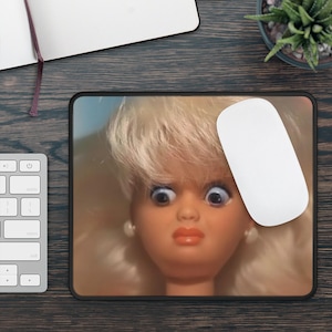 Może przedstawiać: Podkładka pod mysz z zbliżeniem twarzy lalki z szeroko otwartymi oczami i zaskoczoną miną. Lalka ma blond włosy, perłowe kolczyki i czerwoną szminkę. Biała mysz komputerowa spoczywa na podkładce.