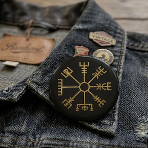 May include: A circular black pin featuring a gold Vegvisir symbol, also known as a Viking compass. The pin is attached to a dark blue denim jacket, which also displays two smaller, separate metal pins near the collar area.