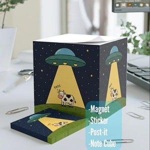 Puede incluir: Un bloc de notas azul oscuro y un imán rectangular a juego con una vaca de dibujos animados siendo abducida por un OVNI verde. El diseño incluye la palabra "Yikes" y un cielo estrellado.