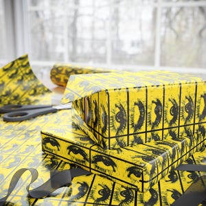 May include: Yellow gift boxes and wrapping paper with a repeating black eye design. A pair of scissors and a black ribbon are also visible. The boxes are stacked on top of each other, ready for gifting.