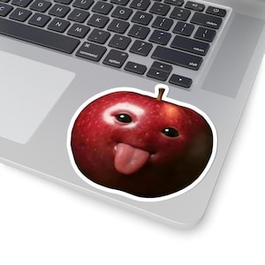 May include: A red apple sticker with a cartoon face, featuring black eyes and a pink tongue sticking out. The sticker is on a laptop, with the keyboard visible.