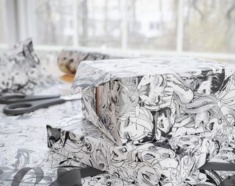May include: Two gift boxes wrapped in black and white manga-style patterned paper. The boxes are stacked on top of each other. A pair of scissors and a roll of matching wrapping paper are visible in the background. A black ribbon is draped across the table.