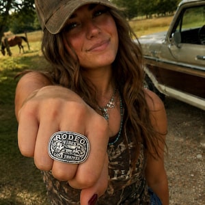 May include: A silver-colored ring with the words "RODEO VIBES LOVE THRIVES" engraved on it. The ring features a design of a horse and rider. The person in the image is wearing a brown cap with the words "LONG LIVE COWGIRLS".