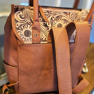 Pode incluir: Mochila de couro castanho com um design floral na aba. A mochila tem al&ccedil;as ajust&aacute;veis e uma al&ccedil;a superior. O design floral &eacute; em relevo e apresenta girass&oacute;is. A bolsa tem um bolso lateral e um fecho de fivela.
