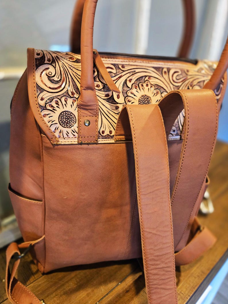 Pode incluir: Mochila de couro castanho com um design floral na aba. A mochila tem al&ccedil;as ajust&aacute;veis e uma al&ccedil;a superior. O design floral &eacute; em relevo e apresenta girass&oacute;is. A bolsa tem um bolso lateral e um fecho de fivela.