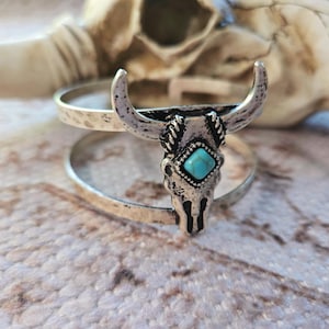 May include: Silver-toned cuff bracelet featuring a bull skull design. The skull has curved horns and a turquoise-colored stone in the center. The bracelet has two bands with a hammered texture, creating a rustic, southwestern aesthetic.