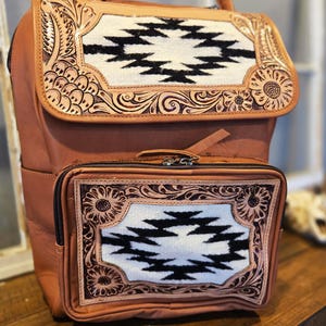 Pode incluir: Mochila de couro castanho com uma al&ccedil;a superior e um bolso frontal. A mochila apresenta um painel decorativo com um design geom&eacute;trico a preto e branco, emoldurado por ferramentas florais e de rolagem.