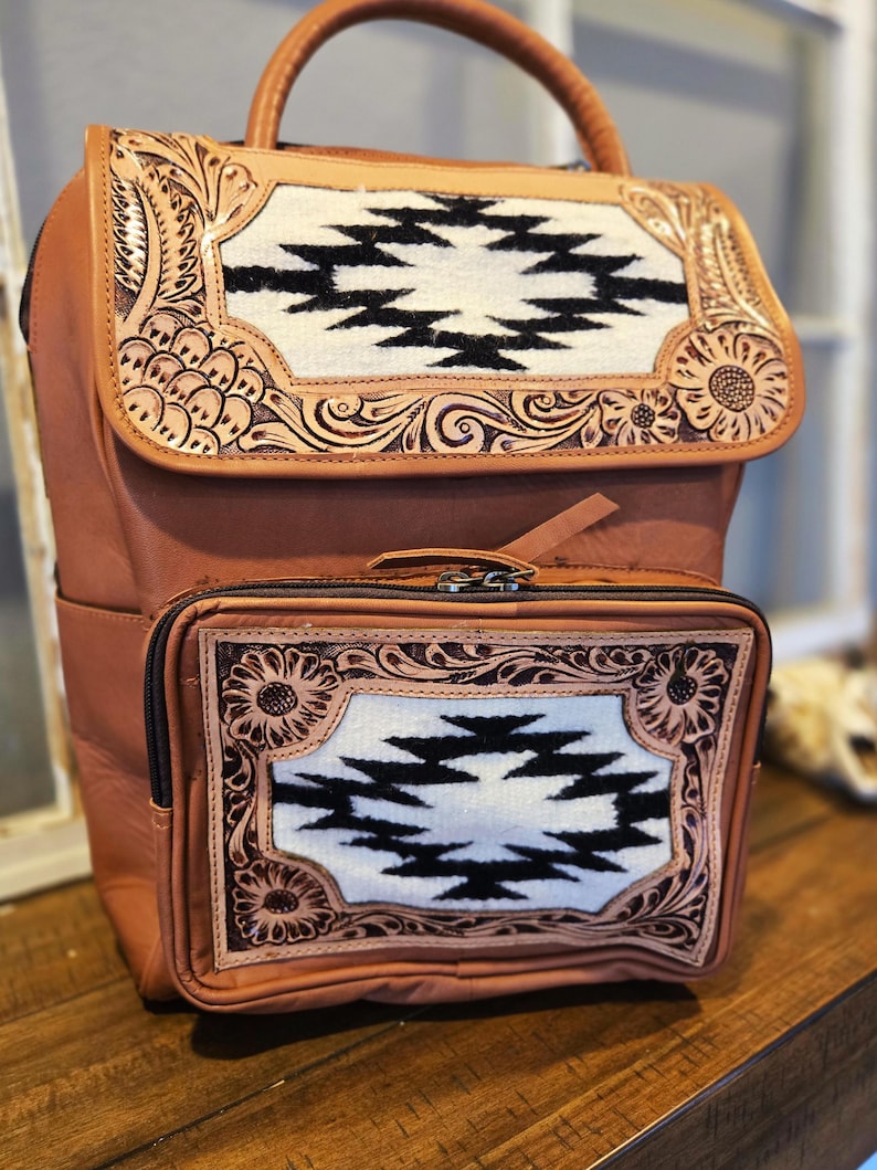 Pode incluir: Mochila de couro castanho com uma al&ccedil;a superior e um bolso frontal. A mochila apresenta um painel decorativo com um design geom&eacute;trico a preto e branco, emoldurado por ferramentas florais e de rolagem.