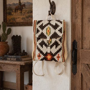 May include: A Southwestern-style backpack with a geometric pattern in brown, white, and orange hangs from a wall hook. The bag features a flap closure and adjustable straps. The background includes a painting and wooden door.