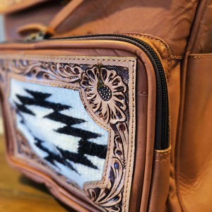 Pode incluir: Close-up de uma mochila de couro castanho com um bolso frontal decorativo. O bolso apresenta um padr&atilde;o geom&eacute;trico branco e preto emoldurado por desenhos florais intrincadamente esculpidos. Um z&iacute;per preto e bolsos adicionais s&atilde;o vis&iacute;veis.