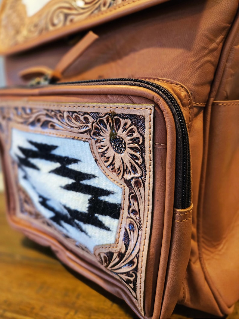Pode incluir: Close-up de uma mochila de couro castanho com um bolso frontal decorativo. O bolso apresenta um padr&atilde;o geom&eacute;trico branco e preto emoldurado por desenhos florais intrincadamente esculpidos. Um z&iacute;per preto e bolsos adicionais s&atilde;o vis&iacute;veis.