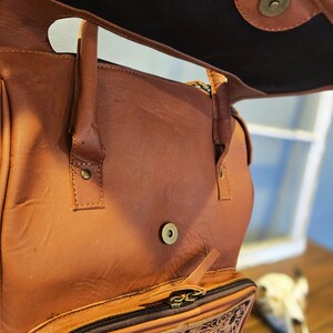 Pode incluir: Mochila de couro marrom com aba, com um bolso frontal com um design floral e geom&eacute;trico decorativo. A bolsa tem uma al&ccedil;a superior e um compartimento com z&iacute;per. O interior &eacute; escuro.
