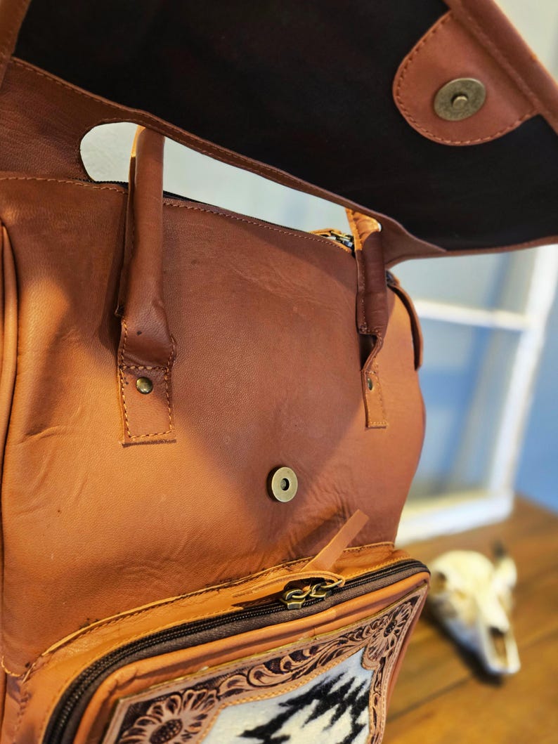 Pode incluir: Mochila de couro marrom com aba, com um bolso frontal com um design floral e geom&eacute;trico decorativo. A bolsa tem uma al&ccedil;a superior e um compartimento com z&iacute;per. O interior &eacute; escuro.