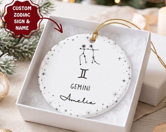 Op de afbeelding: Een rond wit keramieken ornament met het zwarte sterrenbeeld Tweelingen, het Tweelingen-symbool, de tekst Gemini en de naam Amelia. Het ornament is versierd met kleine zwarte stermotieven en hangt aan een gouden koord in een witte doos.