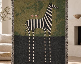 Op de afbeelding: Een geweven plaid met een zwart-witte zebra die op een donkergrijze ondergrond staat tegen een textuurrijke olijfgroene achtergrond. De zebra heeft twee roze cocktailglazen op zijn rug en lange, dunne, gestreepte poten.