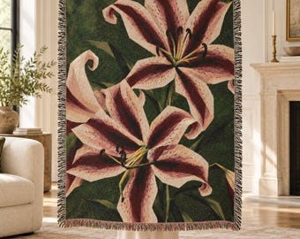 Puede incluir: Una manta tejida que presenta un estampado botánico a gran escala de dos lirios en flor con pétalos de color burdeos oscuro y crema sobre un fondo verde bosque oscuro, acabado con un borde decorativo de flecos en los cuatro lados.