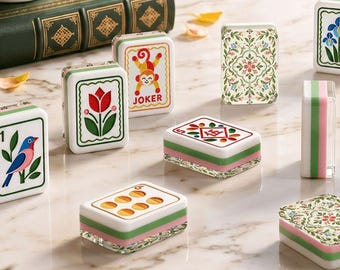 Op de afbeelding: Een set rechthoekige mahjongtegels met diverse kleurrijke illustraties, waaronder een vogel, een tulp, een aap met het label JOKER, bloemmotieven en geometrische ontwerpen. De tegels hebben witte vlakken met groene en roze gelaagde onderkanten.