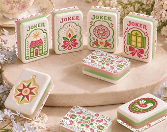 Op de afbeelding: Een verzameling rechthoekige tegels met het woord JOKER en diverse kleurrijke illustraties, waaronder een huis, bloemmotieven, een raam en een vogel. De witte tegels met groene en roze accenten zijn gerangschikt op een ronde stenen ondergrond.
