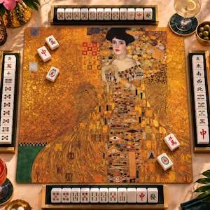 May include: A square mahjong board featuring a gold-toned print of Gustav Klimt's Portrait of Adele Bloch-Bauer I. The set includes four wooden tile racks holding white mahjong tiles with red, green, and black characters, plus loose tiles on the board.